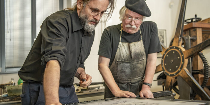 Zwei Männer, beide tragen Brille, stehen an einer historischen Druckmaschine.